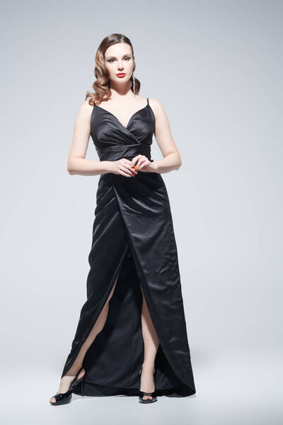 Великолепная девушка-модель с элегантным макияжем и прической позирует в черном вечернем платье. Студийный портрет на белом фоне. Праздничная мода. Портрет в полный рост.