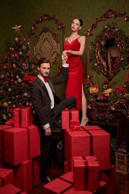 Noel ve yeni yıl modası. Lüks gece elbiseleri içinde yakışıklı bir adam ve güzel bir kız Noel için süslenmiş pahalı iç mekanlarda kırmızı hediye kutuları arasında poz veriyorlar. Tatil indirimi ve alışveriş konsepti. 