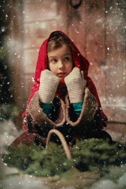Believe in a miracle at Christmas. The little heroine of an old fairy tale sits at a wooden house on Christmas Eve with fir-tree branches in her basket. Magic Christmas night with heavy snowfall. clipart