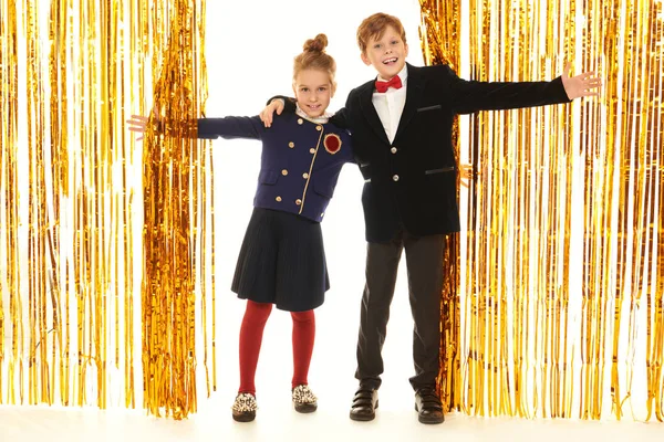 Gece kıyafetleri içindeki neşeli kız ve erkek disko partisindeki parlak altın folyo perdelerin arasında birbirlerini omuzlarından tutuyorlar. Çocuk partisi. Çocuk modası ve tatiller. Tam boy çekim.