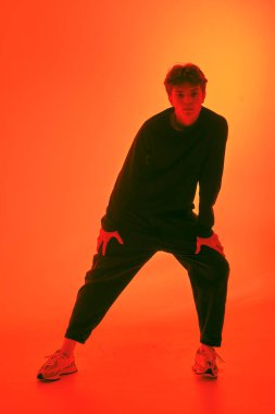 Siyah günlük giysiler ve turuncu arka planda poz veren spor ayakkabıları olan çekici, koyu renk saçlı bir adamın stüdyo fotoğrafı. Tam yol ileri. Gençlik modası ve tarzı.
