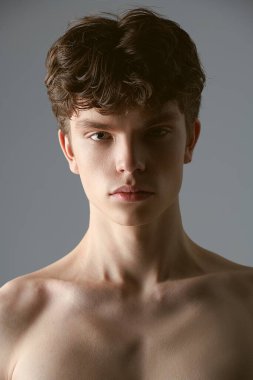 Kahverengi dalgalı saçlı, gri arka planda üstsüz poz veren yakışıklı bir gencin stüdyo portresi. Erkek güzelliği ve sağlığı. 