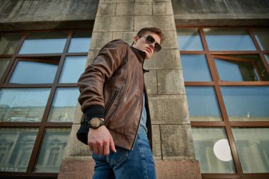 Moda güneş gözlüğü ve deri ceketli kendine güvenen genç adam tuğla bina boyunca bir şehir caddesinde yürüyor. Erkek yaşam tarzı ve moda.