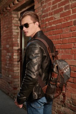 Güneş gözlüklü, deri ceketli, sırtında sırt çantası olan yakışıklı bir adam şehir caddesinde yürüyor. Yaşam tarzı ve erkek modası. 