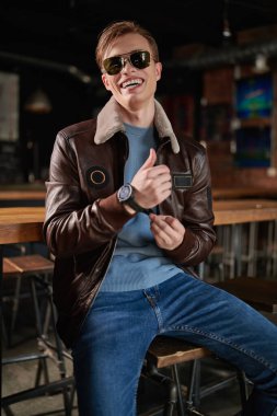 Şık kıyafetler ve güneş gözlüğü takan neşeli sarışın genç adam kol saatini neşeyle bir barda otururken bağlıyor. Erkek modası. Yaşam biçimi. 