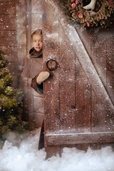 Kışın ortaçağ giysileri içinde, karla kaplı ahşap evin kapısından dışarı bakan sevimli küçük bir kızın portresi. Tarihi yeniden yapılandırma. Noel beklentisi.