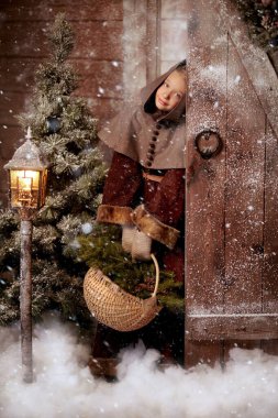 Full length shot of a joyful girl in medieval clothes standing at a door of a snow-covered wooden house with a basket of fir branches and fir cones. Historical reconstruction. Merry Christmas! clipart