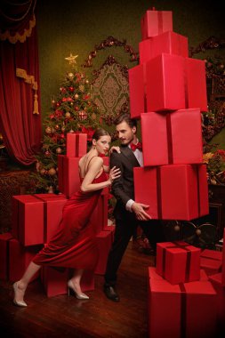 Güzel bir çift şık bir gece elbisesi içinde bir erkek ve bir kadın lüks Noel süslemeleri içinde kırmızı hediye kutuları taşıyorlar. Noel ve Yeni Yıl indirimi, alışveriş.  