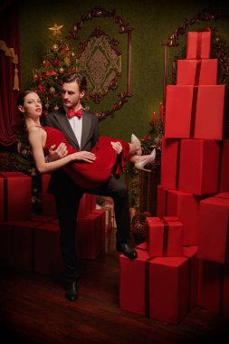 Noel ve Yeni Yıl kutlaması. Zarif bir gece elbisesi içinde yakışıklı bir adam lüks bir Noel 'de duran kırmızı elbiseli sevgilisini kollarında tutuyor. Tatil modası. 