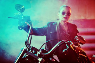 Siyah deri ceketli, siyah güneş gözlüklü kendine güvenen motorcu kız garajda motosikletinin üstünde poz veriyor..