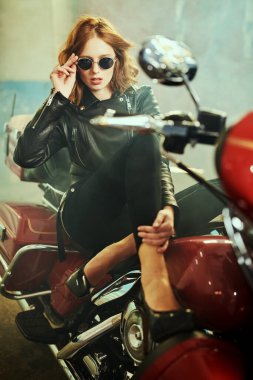 Siyah deri ceketli, güneş gözlüklü kızıl saçlı güzel bir kız kırmızı bir motosiklette oturuyor. Garaj içi..