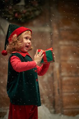 Noel Masalı. Küçük bir elf kızı portresi, tatlı kıvırcık saçlı, kar yağışı altında ahşap bir kulübede duruyor, elinde bir hediye tutuyor ve gülüyor.. 