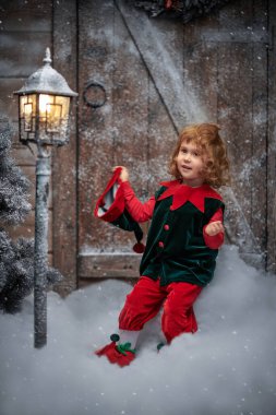 Kış Noel Masalı. Küçük sevimli bir elf kızı güler ve sevinir, kar yığınlarının arasında tahta bir kulübenin yanında durur ve karla kaplı bir fener kar altında kalır..