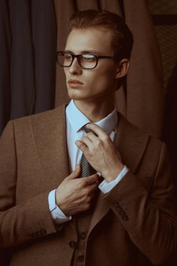 Zarif kahverengi takım elbiseli ve gözlüklü yakışıklı sarışın bir adam lüks klasik takım elbisesinin arka planında kravatını düzeltiyor. Erkeklerin iş modası.