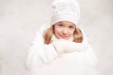 Ilık beyaz örgü elbiseli neşeli küçük kız karda oturmuş büyük bir kartopuna yaslanıyor. Kış çocuklarının moda ve aktiviteleri.