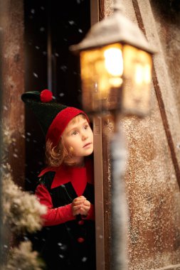 Kış Noel Masalı. Tatlı küçük elf kızı, muhteşem bir kış ormanında, karla kaplı ahşap bir kulübenin kapısından dışarı bakar..