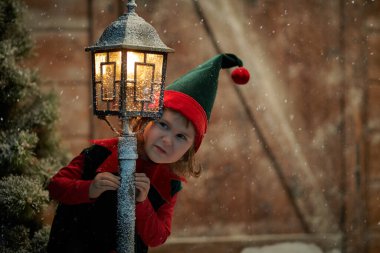 Küçük sevimli bir elf kızı kar taneleri yakalar ve sevinir. Tahta bir kulübede ve kar yağışı altında karla kaplı bir fenerde durur. Kış Noel Masalı.