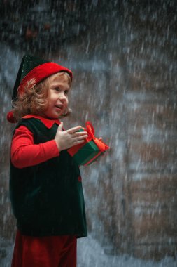 Noel Masalı. Küçük bir elf kızı portresi, tatlı kıvırcık saçlı, kar yağışı altında ahşap bir kulübede duruyor, elinde bir hediye tutuyor ve gülüyor.. 