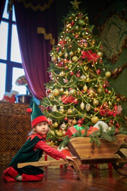 Noel hikayeleri. Şirin küçük kız elinde hediyelerle ahşap bir arabanın yanında duruyor, oyuncağını tutuyor ve gülümsüyor. Şöminesi ve köknar ağacı olan eski bir oda. Noel için çok güzel dekore edilmiş.. 