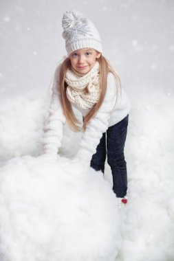 Kış çocuğu modası. Kardan adam yapan, kalın örgü elbiseli neşeli bir kızın portresi. Beyaz karlı arka plan. Kış aktiviteleri ve tatiller. Stüdyo çekimi.