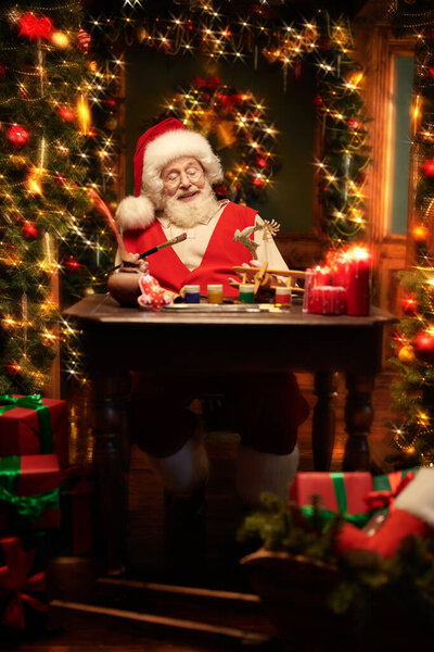 Портрет веселого Санта-Клауса за работой в мастерской по изготовлению и покраске игрушек для подарков детям. Красивая комната празднично украшена в честь Рождества. Концепция Рождества и Нового года. 