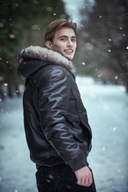 Çekici, siyah deri ceketli, kürklü, çekici sarışın bir adam kış parkında karla kaplı bir şekilde yürürken yüzünde mutlu bir gülümsemeyle geri dönüyor. Erkekler kış modası.