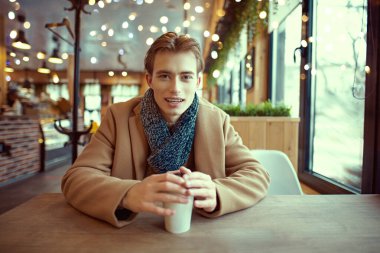 Güzel görünümlü genç bir adamın portresi şık bir palto ve eşarp ile sıcak bir kış gününde sıcak bir kafede ısınıyor ve karşısındaki biriyle konuşuyor..