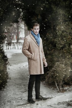 Paltosu ve atkısı olan yakışıklı sarışın genç adam parkta karla kaplı duruyor. Tam yol ileri. Erkek modası ve yaşam tarzı.