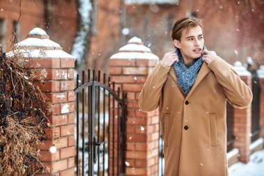 Şık ceketli ve atkılı yakışıklı bir gencin portresi tuğla bir evin kapısında duruyor atkıyı ayarlıyor ve başka tarafa bakıyor. Erkek güzelliği ve modası. Boşluğu kopyala.