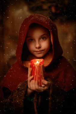 An angelic little girl in medieval clothes with a hood stands under snow by a wooden house with a burning candle carolling and waiting for a Christmas miracle. Believe in a miracle at Christmas.  clipart