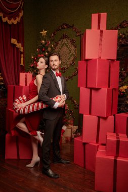 Zarif giyimli genç çift, büyük bir baston şeker tutarak Noel için pahalı iç dekorasyonlarla süslenmiş kırmızı hediye kutularıyla sırt sırta poz veriyor. Noel ve yeni yıl modası ve alışverişi.