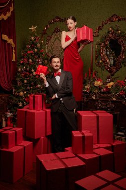 Tatil indirimi ve alışveriş. Lüks gece elbiseleri içinde yakışıklı bir adam ve güzel bir kız Noel için süslenmiş pahalı iç dekorasyonlarda kırmızı hediye kutuları arasında poz veriyorlar. Noel ve Yeni Yıl modası. 