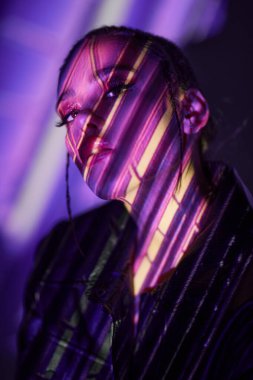 Gelecekçi stil, güzellik ve moda. Siyah deri ceketli çekici esmer kız yüzünde neon ışıklı ışıklarla karanlık bir alanda poz veriyor. Sanal gerçeklik kavramı. 