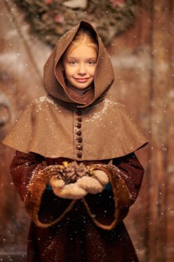 Portrait of a lovely little girl in medieval clothes standing by a wooden house on a snowy day holding fir cones. Historical reconstruction. Christmas in Medieval Times. clipart