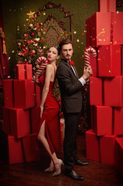 Zarif genç çift, Noel için süslenmiş lüks şeker kamışlarıyla sırt sırta duran kırmızı hediye kutularının arasında. Noel ve yeni yıl modası. Tatil satışı ve alışveriş. 
