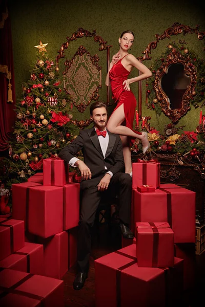 Tatilde alışveriş ve satış. Lüks gece elbiseleri içinde yakışıklı bir genç adam ve güzel bir kız Noel için süslenmiş pahalı iç dekorasyonlarda kırmızı hediye kutularının arasında duruyor. Tatil modası. 