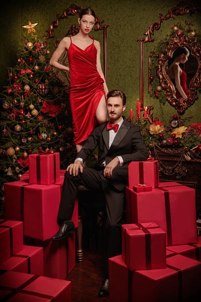 Noel ve yeni yıl modası. Lüks gece elbiseleri içinde yakışıklı bir adam ve güzel bir kız Noel için süslenmiş pahalı iç dekorasyonlarda kırmızı hediye kutularının arasında poz veriyorlar. Tatil indirimi ve alışveriş konsepti. 