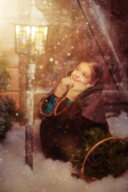 Ortaçağ kıyafetleri içinde şirin küçük bir kız yanan bir fenerin güzelliğine hayran olan bir sepet ladin dallarıyla karla kaplı ahşap bir evde oturuyor. Eski Noel Masalı. 