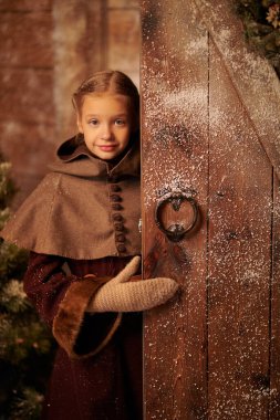 Kışın ortaçağ giysileri içinde, kar kaplı ahşap bir evin kapısından dışarı bakan sevimli küçük bir kızın portresi. Tarihi yeniden yapılandırma. Noel mucizeleri ve beklentileri.