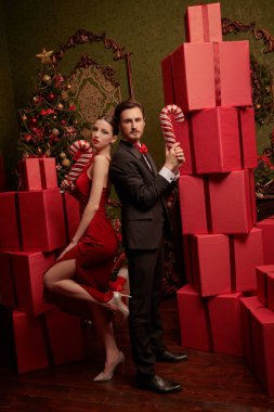 Zarif giyimli genç çift, büyük şeker kamışları tutarak Noel için pahalı iç dekorasyonlarla süslenmiş kırmızı hediye kutularıyla sırt sırta poz veriyor. Noel ve yeni yıl modası ve alışverişi.