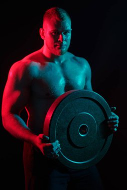 Elinde büyük bir demir disk tutan kaslı yakışıklı bir atletin portresi. Kırmızı ışıklandırmalı koyu bir arka plan. Spor konsepti, halter, vücut geliştirme.. 