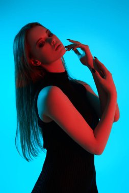 Güzellik ve moda portresi. Siyah dumanlı göz makyajı ve uzun düz saçlarıyla mavi arka planda kırmızı ışıkta siyah dar bir elbise giymiş güzel bir kız. Makyaj ve kozmetik.