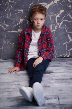 Çocuk modası. Giyim kuşam giyinmiş yakışıklı bir çocuğun uzun bir portresi. Yerde grunge duvarın arkasında oturuyor.. 