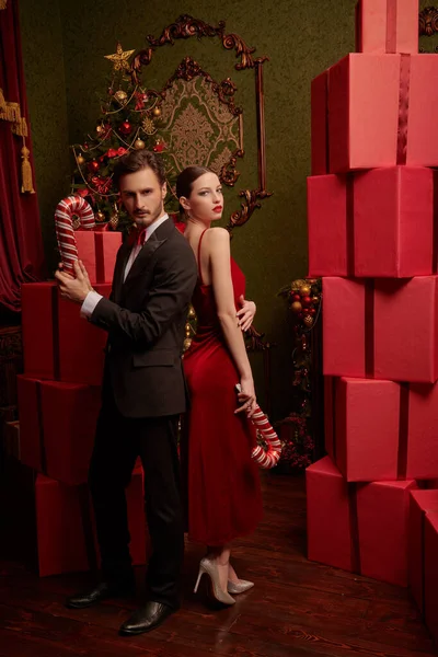 Zarif gece kıyafetleri giymiş genç bir çift Noel için dekore edilmiş lüks iç dekorasyonlarda kırmızı hediye kutularının yanında sırt sırta verip duruyor. Noel ve yeni yıl modası ve alışverişi.