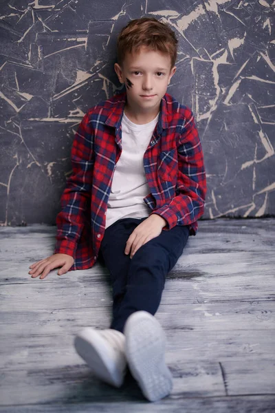 Çocuk modası. Giyim kuşam giyinmiş yakışıklı bir çocuğun uzun bir portresi. Yerde grunge duvarın arkasında oturuyor.. 