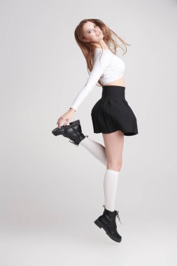 Youth style. Full length shot of pretty, sexy girl in stylish white top, black school skirt, socks and boots jumping joyfully on a white studio background. Modern youth dances. 