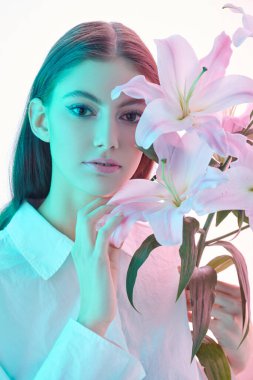 Beauty and flowers. Portrait of a beautiful brown-haired girl with a delicate lily flower on a glowing white background. Make-up and cosmetics. Perfumery. 