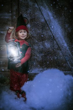 Magical Christmas night. A portrait of a cute smiling little elf girl standing in the snowy forest by a wooden house holding joyfully a burning lantern. Miracles for Christmas.
