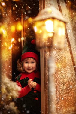 Christmas fairy tale. Cute little elf girl looks out from behind the door of a snow-covered wooden hut in a fabulous winter forest.