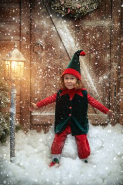 A little cute elf girl smiles and rejoices, sitting on a big snowball near a wooden house and a snow-covered lantern under a snowfall. Christmas fairy tales. 